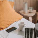 Pen Diary - From above of open netbook with black screen and cup of coffee with smartphone and notebook placed on crumpled bed sheet in bedroom with big vase on floor