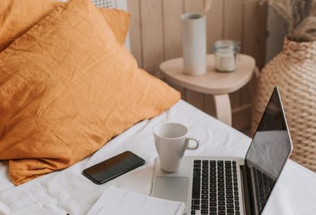 Pen Diary - From above of open netbook with black screen and cup of coffee with smartphone and notebook placed on crumpled bed sheet in bedroom with big vase on floor