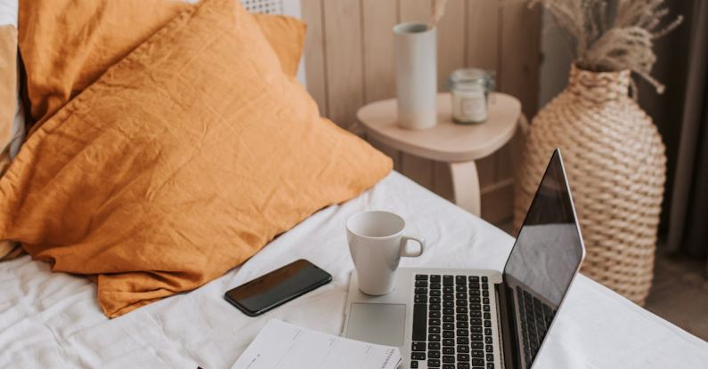 Pen Diary - From above of open netbook with black screen and cup of coffee with smartphone and notebook placed on crumpled bed sheet in bedroom with big vase on floor