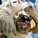 Earth Tech - This picture shows an american astronaut in his space and extravehicular activity suite working outside of a spacecraft. In the background parts of a space shuttle are visible. In the far background of the picture planet earth with it's blue color and white clouds is shown as well as a patch of black space.