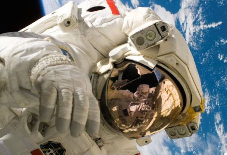 Earth Tech - This picture shows an american astronaut in his space and extravehicular activity suite working outside of a spacecraft. In the background parts of a space shuttle are visible. In the far background of the picture planet earth with it's blue color and white clouds is shown as well as a patch of black space.