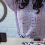 Phone Mirror - Stylish young African American female influencer in sweater adjusting long dark hair while sitting at wooden table before recording makeup tutorial video on smartphone