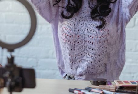 Phone Mirror - Stylish young African American female influencer in sweater adjusting long dark hair while sitting at wooden table before recording makeup tutorial video on smartphone