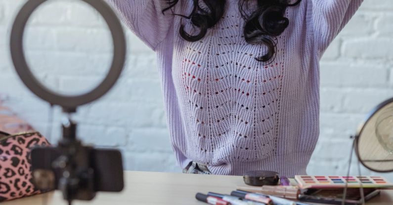 Phone Mirror - Stylish young African American female influencer in sweater adjusting long dark hair while sitting at wooden table before recording makeup tutorial video on smartphone